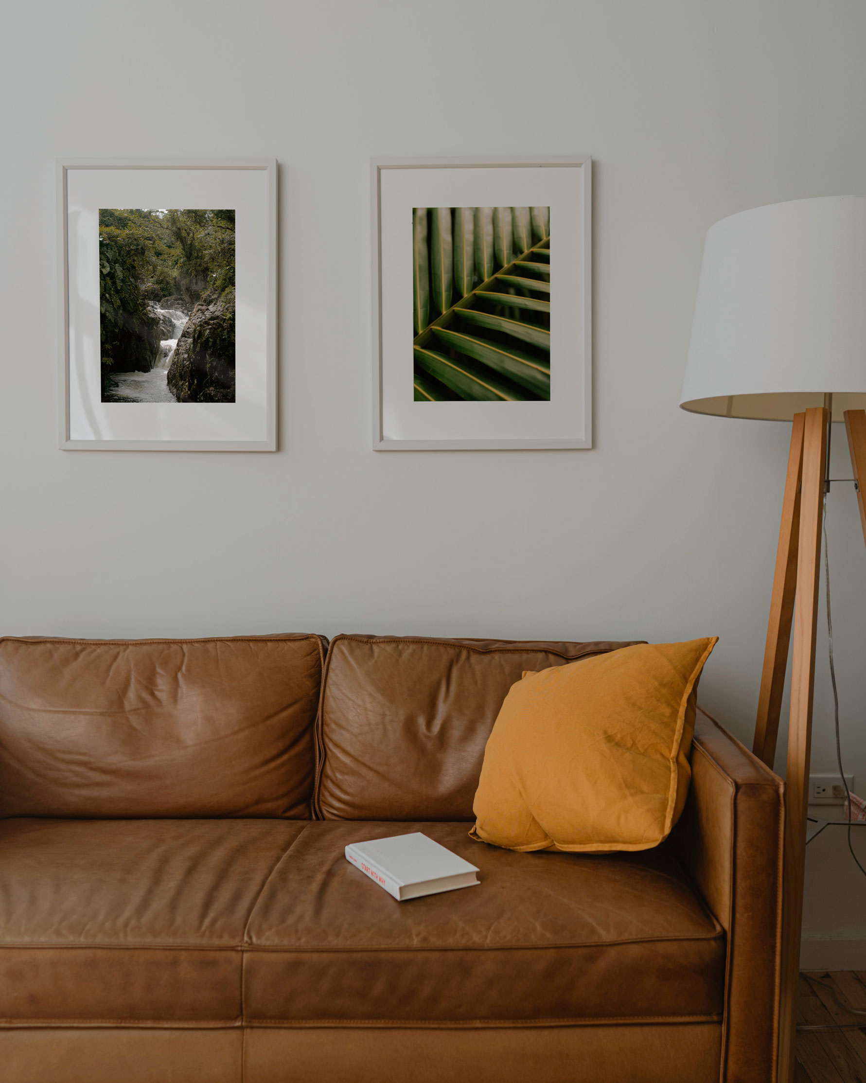 A cozy living room setting with a brown leather couch, a yellow throw pillow, a white book on the couch, and two framed nature photographs hanging on the wall.