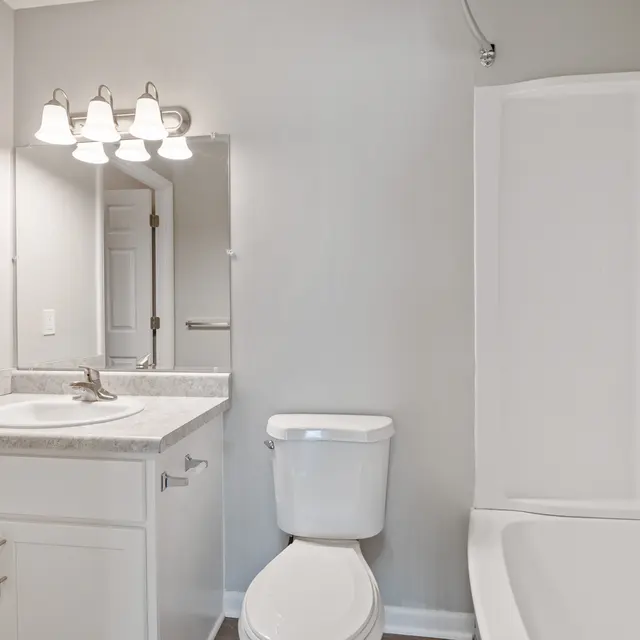 A clean and modern bathroom featuring a sink with a countertop, a toilet, and a bathtub with a shower. The walls are painted in a light gray tone and there are three light fixtures above the mirror.