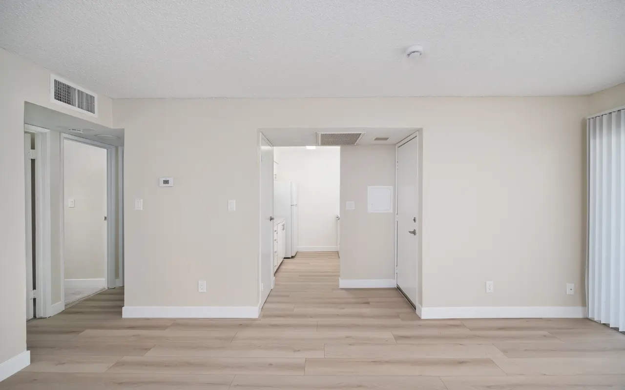An interior view of an apartment showing a spacious room with light-colored walls and wood flooring. Doorways lead to other rooms, with a glimpse of a kitchen in the background.