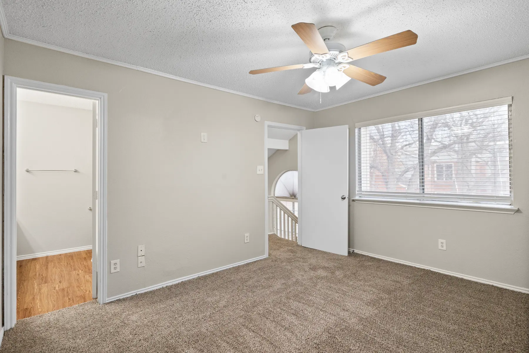 A well-lit, empty bedroom featuring light gray walls, a ceiling fan with wooden blades, and a window with a view of bare trees. The room has plush carpeting and a door leading to a small staircase.