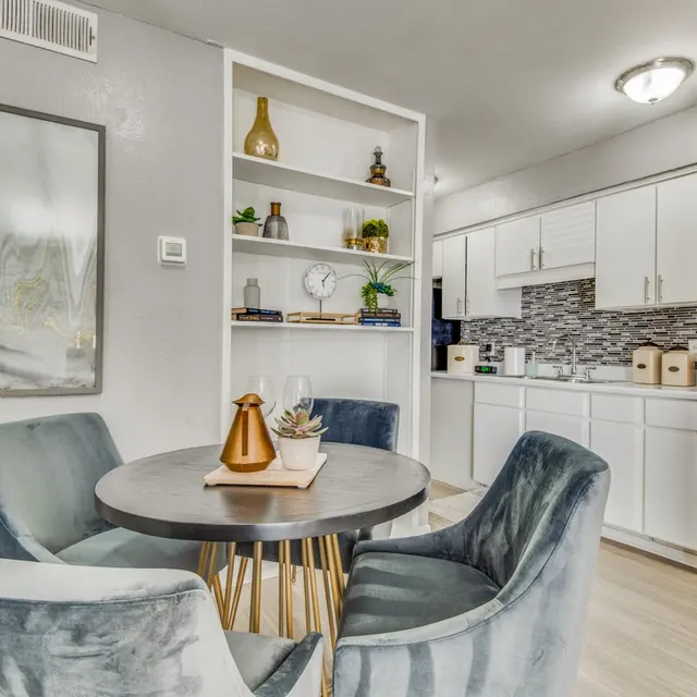 A modern dining area featuring a circular table surrounded by four plush chairs. There is a decorative centerpiece on the table and a white shelving unit in the background filled with decorative items. The kitchen area is visible, featuring white cabinets and a tiled backsplash.