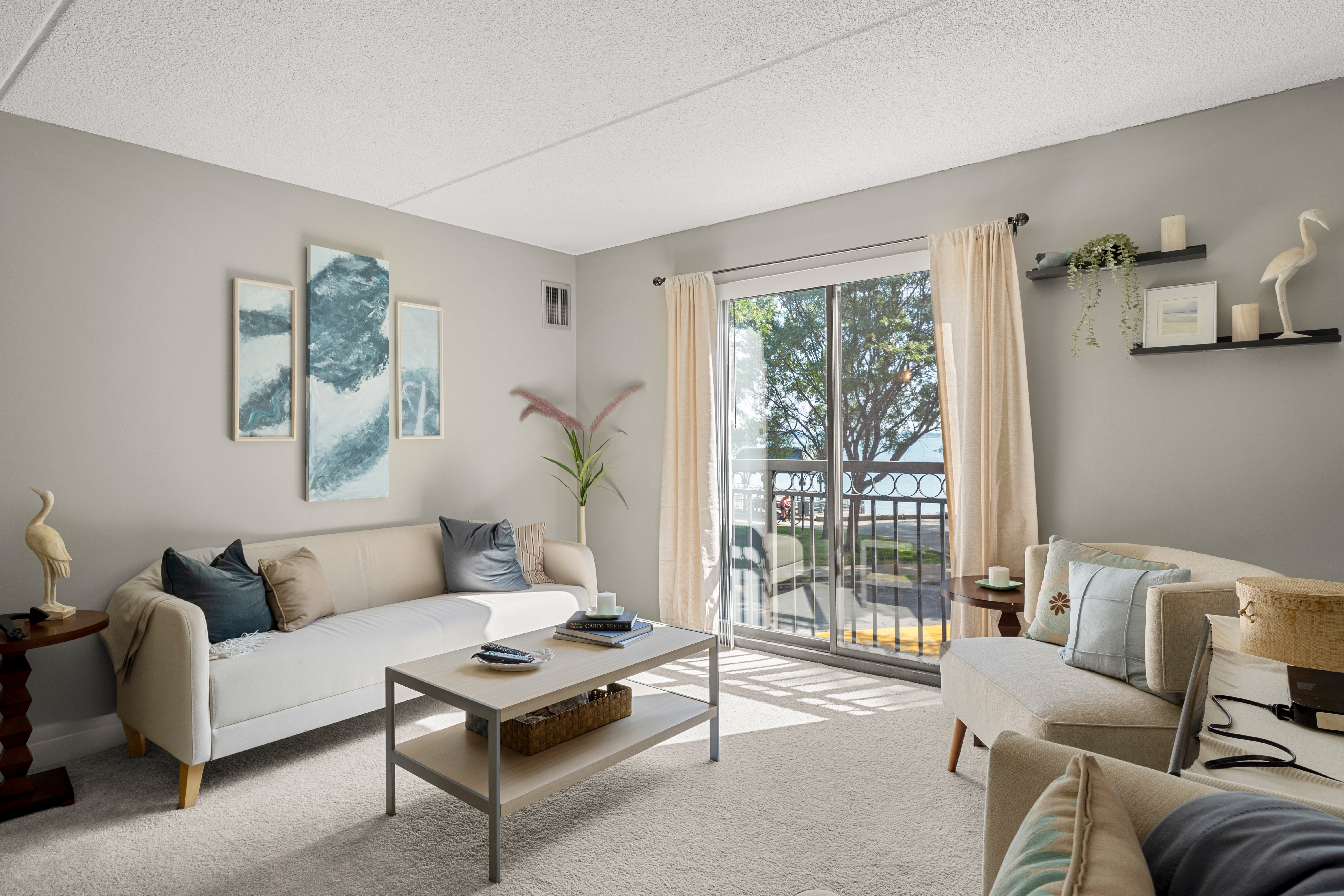 Cozy living room featuring a white sofa, small coffee table, and large windows with natural light. Decor includes wall art, potted plants, and accent pillows on the sofa.