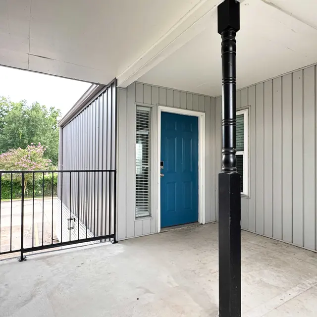 A covered porch area with a blue door and modern gray siding. There is a railing and views of greenery in the background.