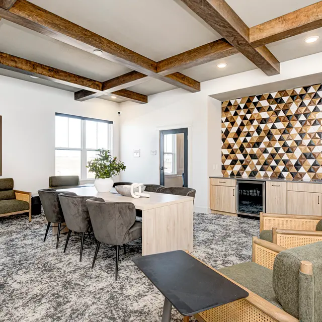 A modern lounge area featuring a large rectangular table surrounded by upholstered chairs, with two armchairs on the side. The room has a decorative wall with geometric patterns and wooden ceiling beams, complemented by a light-colored carpet.