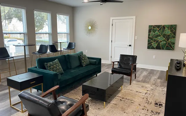A modern living room featuring a teal couch with decorative pillows, two black leather chairs, a glass coffee table, and a patterned rug. The room has large windows allowing natural light and minimalistic decor with a wall art piece and a decorative wall feature.