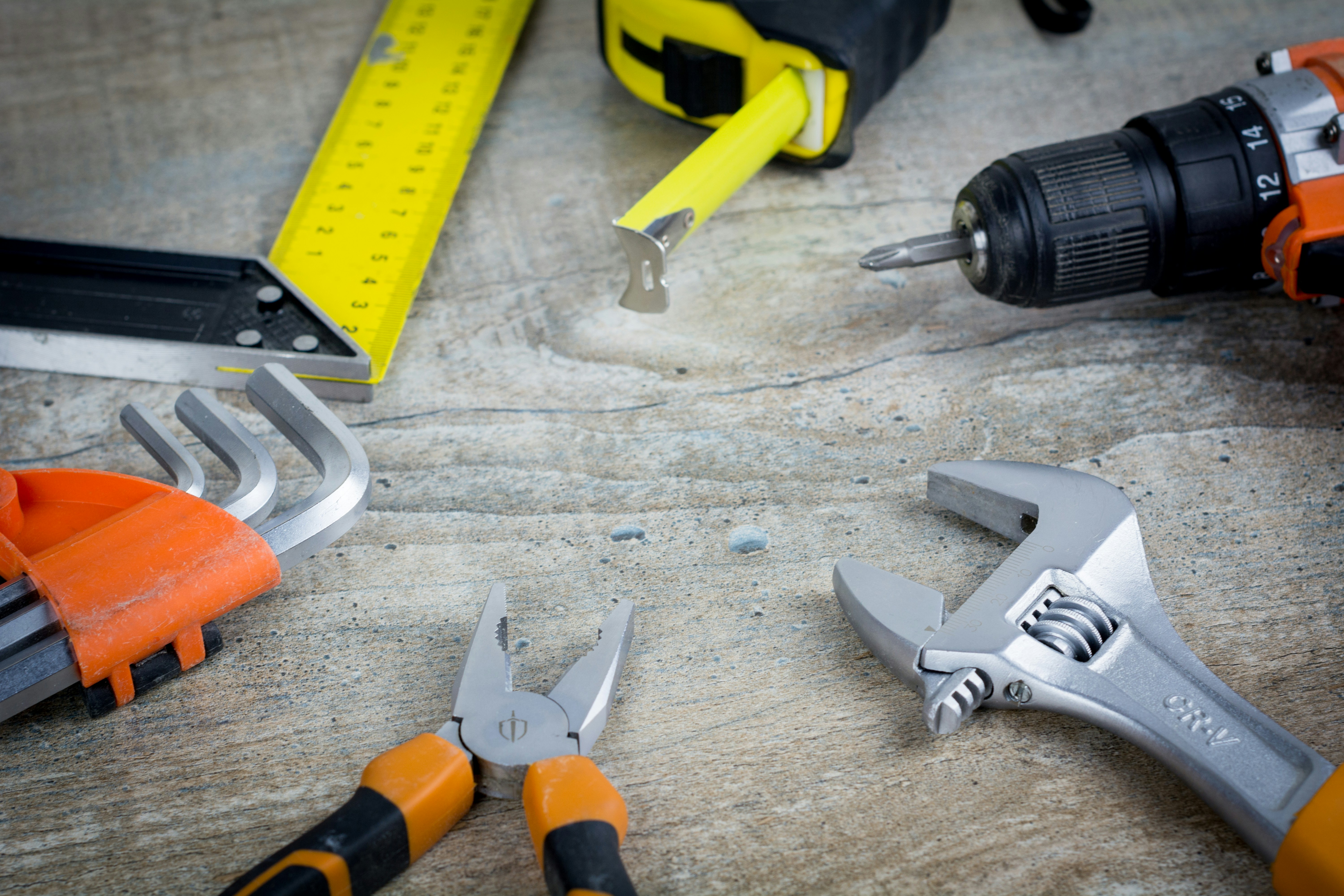 A collection of various tools laid out on a wooden surface. The tools include a tape measure, a drill, a wrench, pliers, and a set of hex keys.