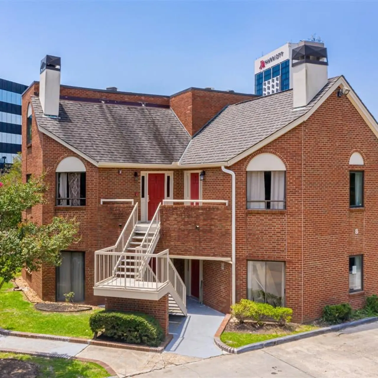 A two-story brick apartment building with a central staircase, surrounded by greenery. In the background, a high-rise building is visible.
