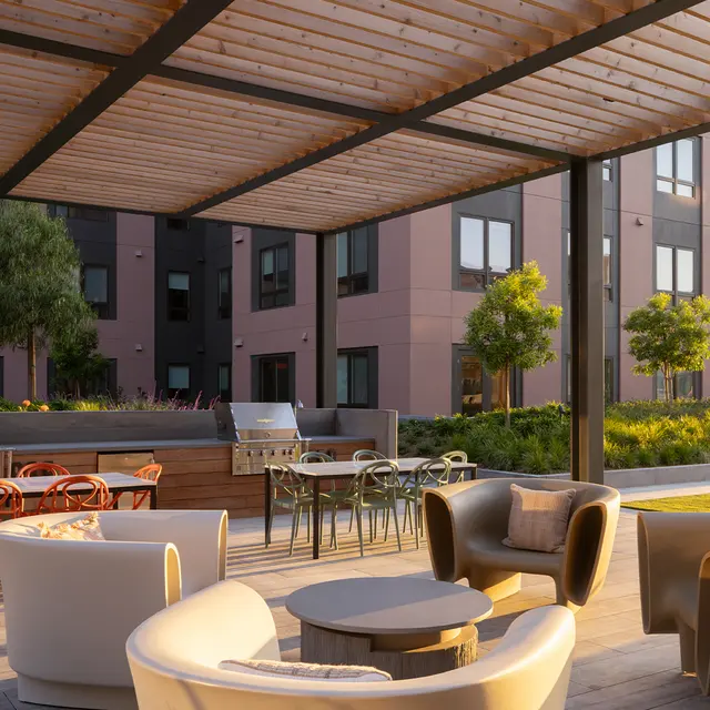 Modern outdoor lounge area with stylish furniture including armchairs, a circular table, and wooden dining tables under a shaded pergola, surrounded by greenery and modern architecture.