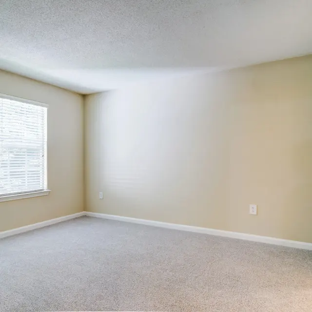 A vacant room with neutral-colored walls and carpet, featuring a window with blinds on one side and an open door on the right.