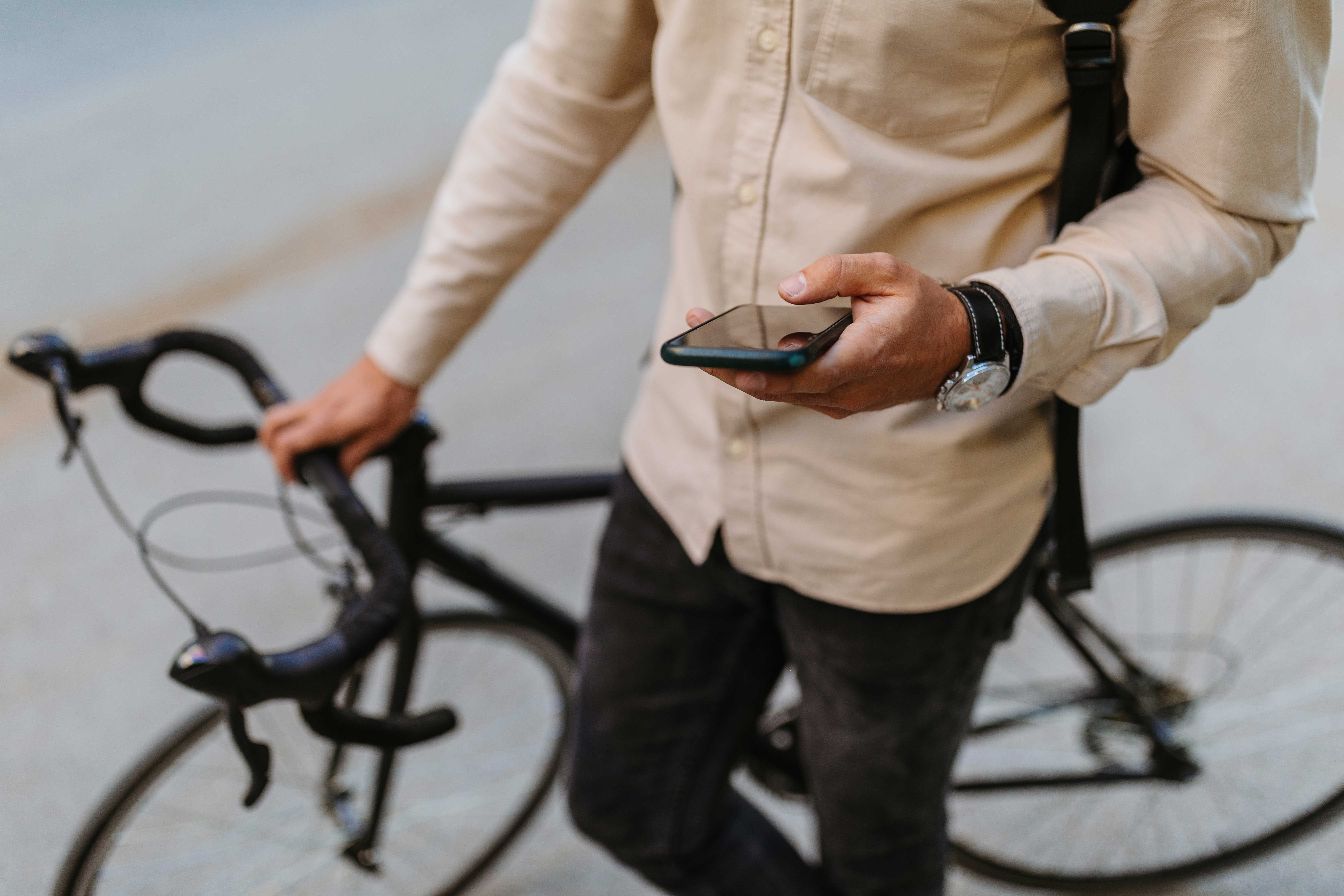 A person holding a smartphone while standing next to a bicycle. The individual is wearing a light-colored button-up shirt and dark pants, with a watch on their wrist. The bicycle has a sleek design and is positioned to the side.
