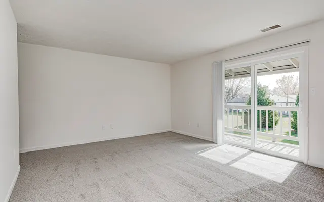 A spacious, empty living room with beige carpet and white walls. A sliding glass door leads to a balcony with a view of greenery outside. Natural light fills the room from the door.