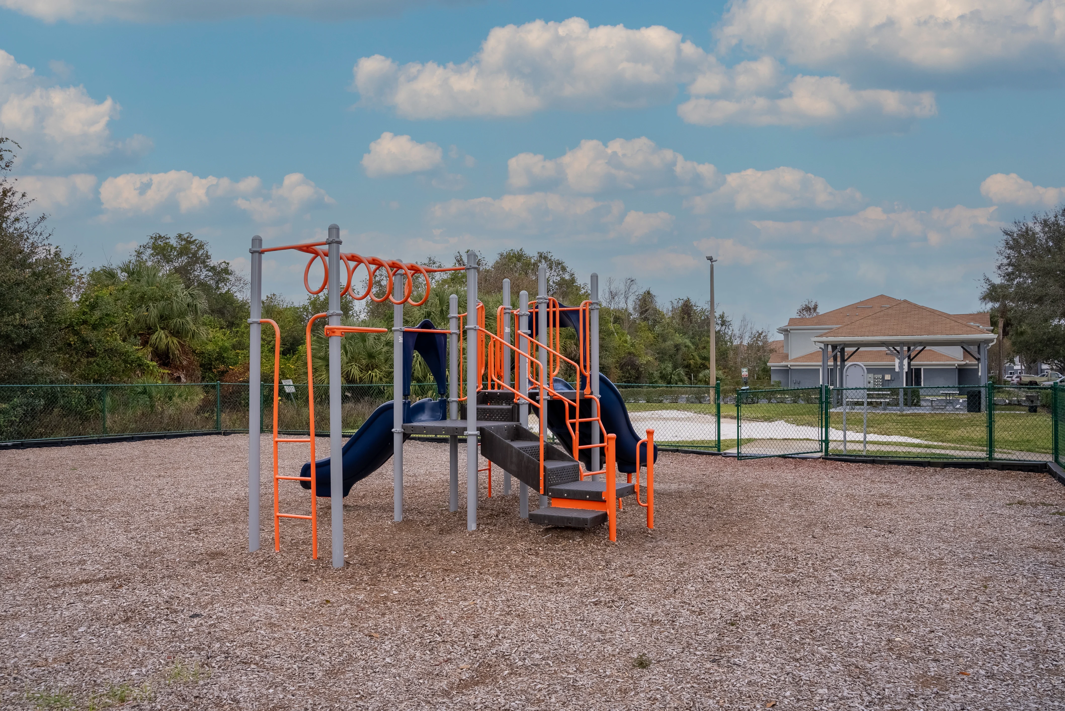 A playground structure with a slide and monkey bars in a gravel area, surrounded by grass and trees, with a building in the background.