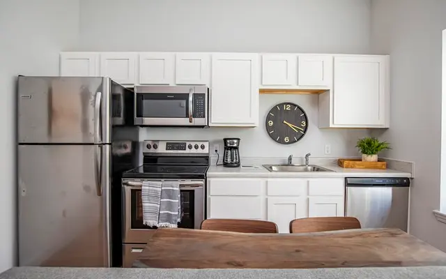 A modern kitchen with stainless steel appliances including a refrigerator, microwave, and oven. The kitchen features white cabinets, a wooden countertop, and a clock on the wall. There is a coffee maker on the counter and a plant adding a touch of greenery.