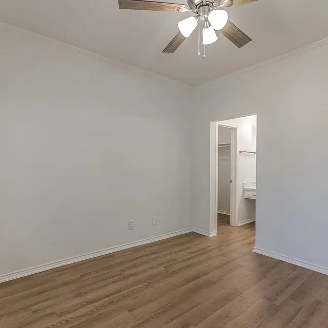 A bright, empty room featuring light-colored walls and wooden flooring. A ceiling fan with lights hangs from the ceiling, and a doorway is visible leading to another space.