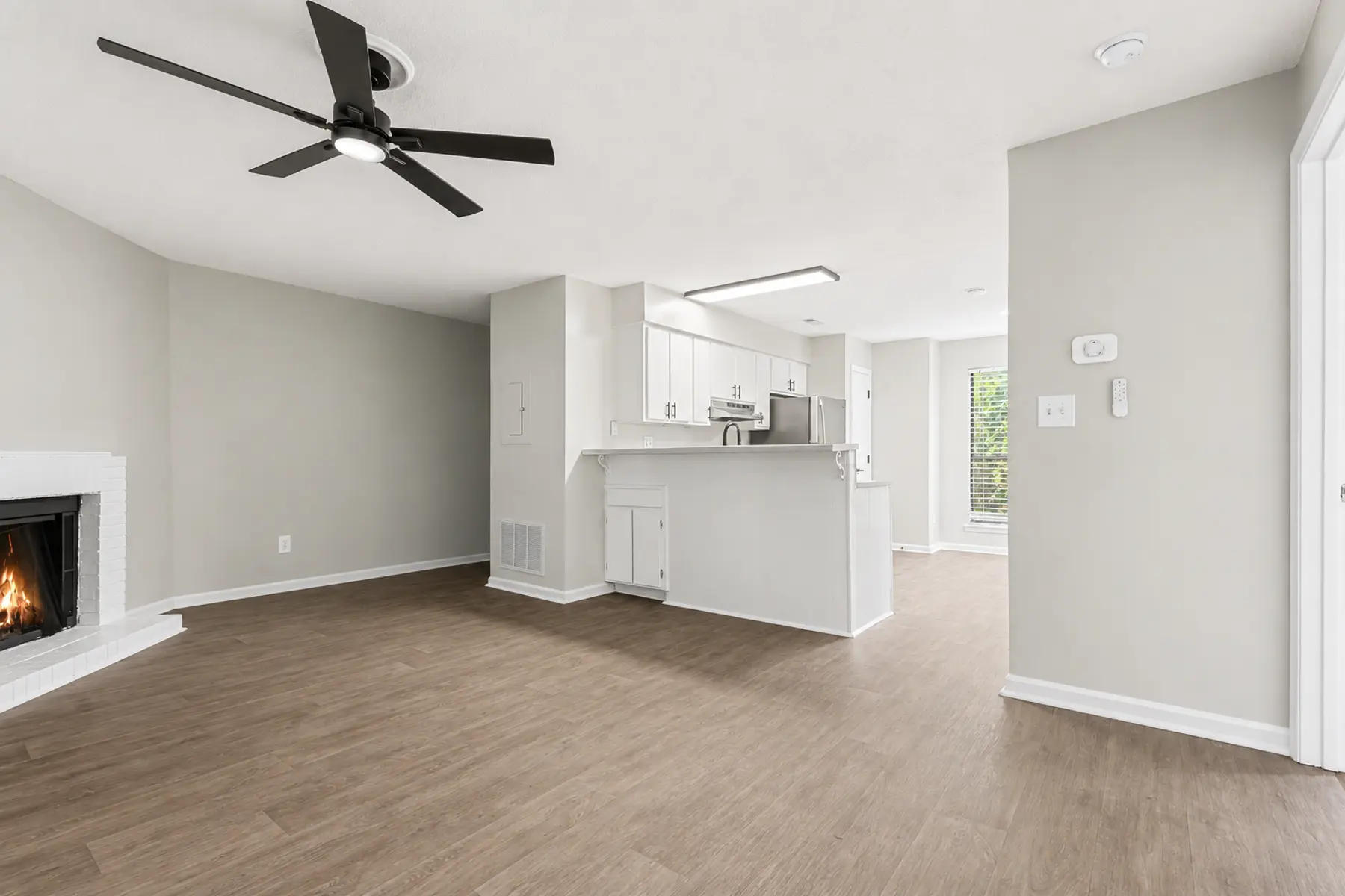 A modern apartment interior showcasing an open living room area with wood flooring and a kitchen island. There is a ceiling fan, and a small fireplace on the left side. Natural light is coming from large windows.