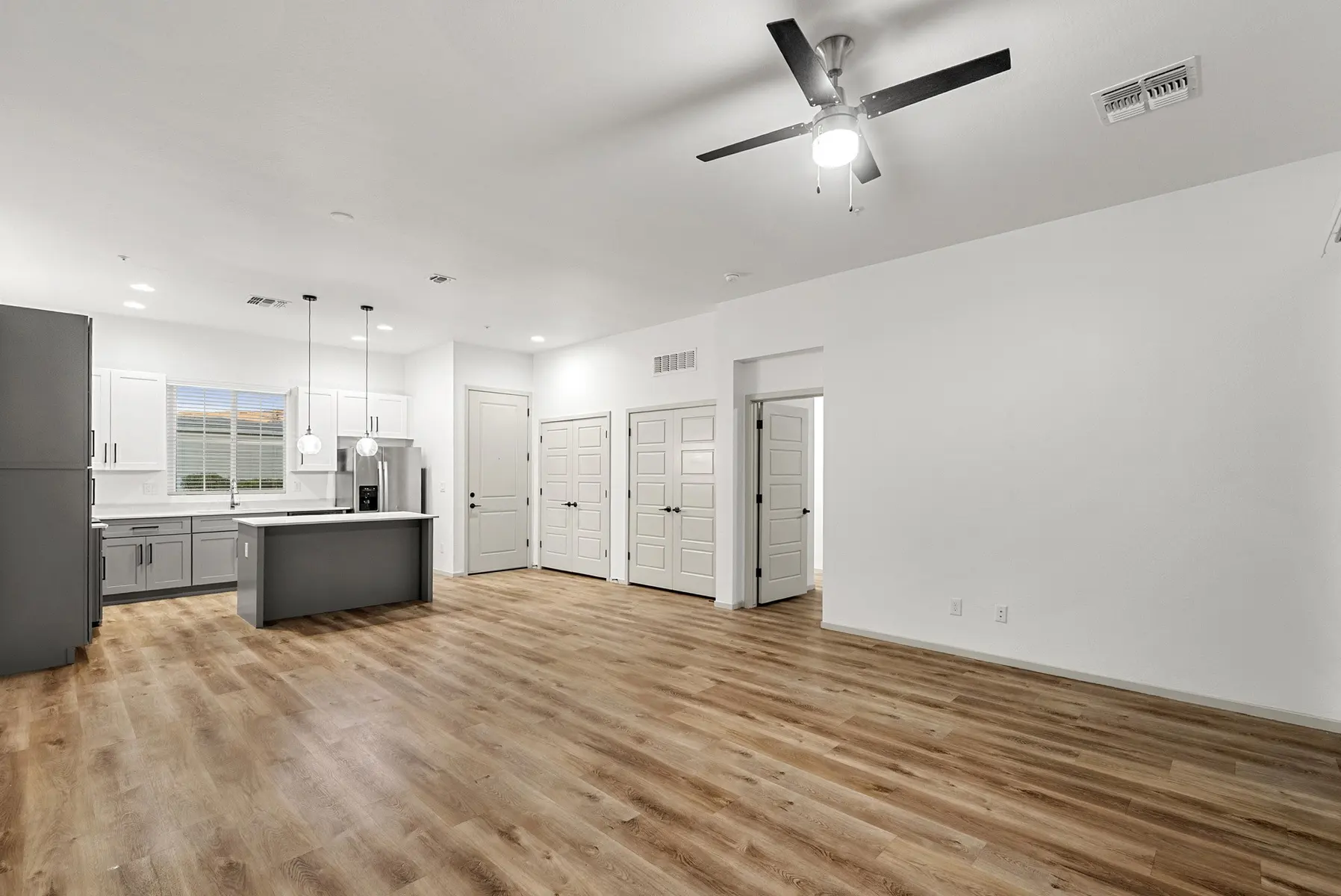 Modern open-concept living area with wood flooring, a kitchen island, and white walls, featuring multiple doors and a ceiling fan.