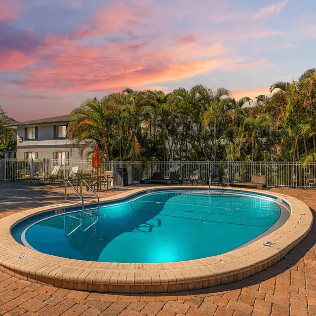 A serene swimming pool with clear blue water surrounded by palm trees at sunset, with lounge chairs on a brick patio nearby.