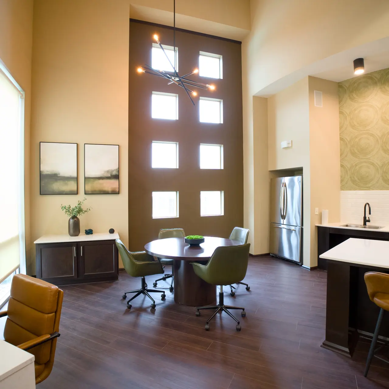 A modern office space featuring a high ceiling with large windows, a round table surrounded by green chairs, a white countertop, and stylish decor.