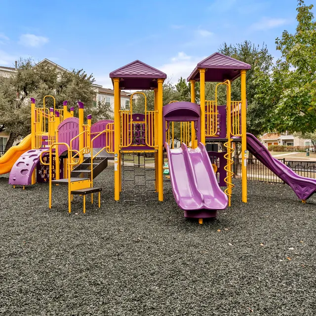 A colorful playground featuring yellow and purple slides, climbing structures, and safety barriers surrounded by trees.