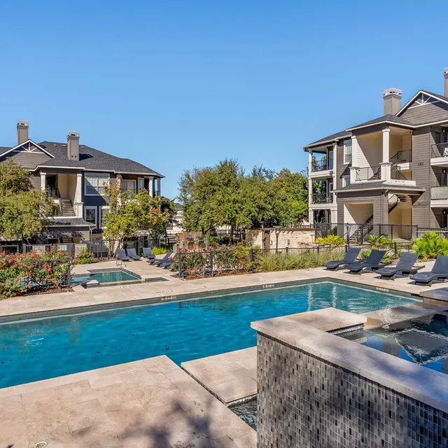 View of a swimming pool surrounded by landscaped areas and apartment buildings on either side, featuring lounge chairs and clear blue sky.
