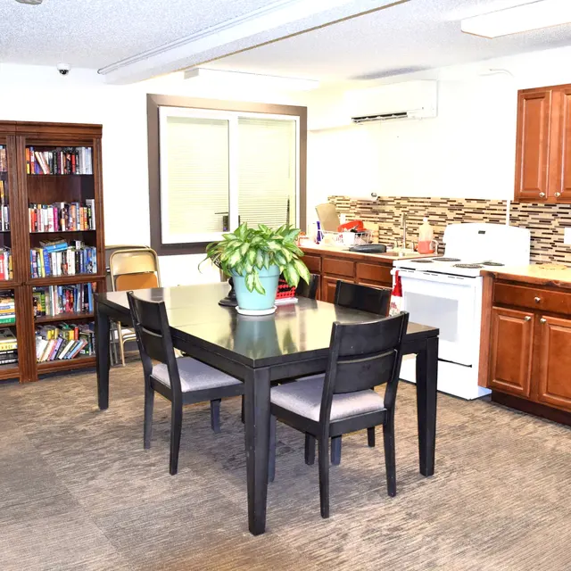 Cozy Kitchen and Dining Area An interior view of a kitchen and dining area featuring a wooden table with four chairs, a potted plant in the center, and a nearby bookshelf filled with books. The kitchen has wooden cabinets and modern appliances.