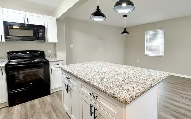 Modern kitchen featuring a central island, dark appliances, and pendant lights, with an open living space in the background.
