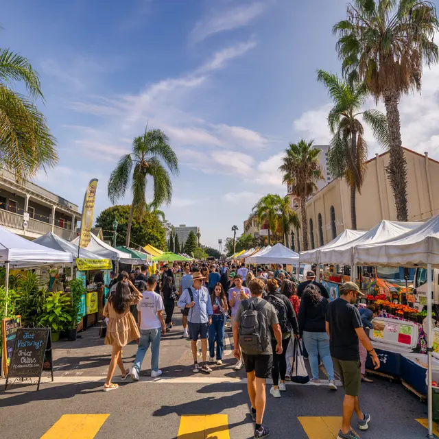 A bustling farmers market with people walking among white tents and palm trees under a blue sky.