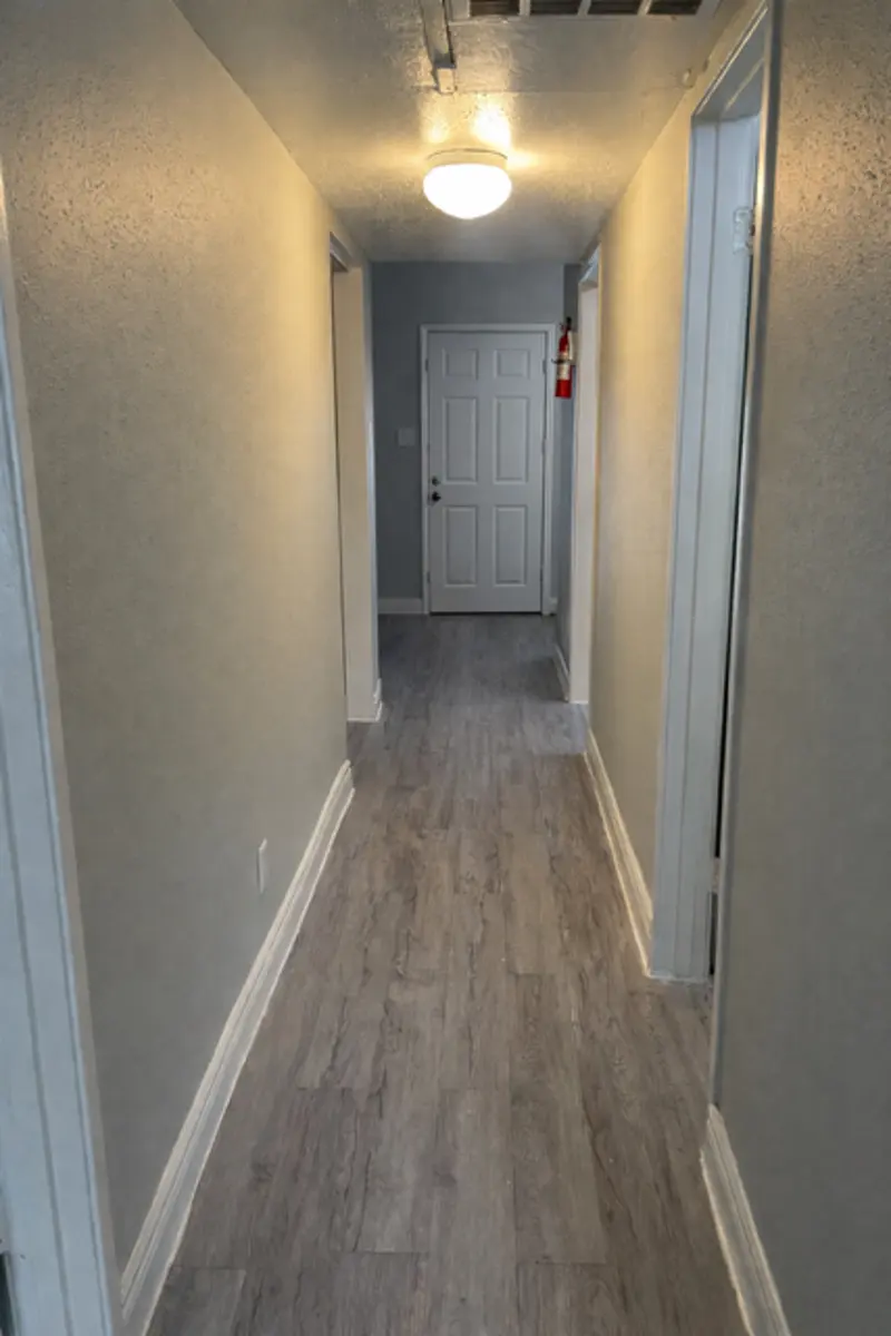 A well-lit hallway with light gray walls and wooden flooring. A closed white door is visible at the end of the hallway.