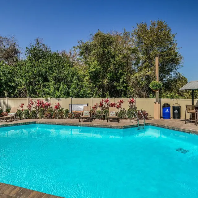 A bright and inviting swimming pool area surrounded by lush greenery and colorful flowers. The pool features clear blue water and is bordered by lounge chairs under the sun. A shaded seating area with a barbecue station is in the background.