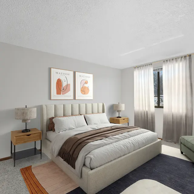 A cozy modern bedroom featuring a queen-sized bed with neutral bedding, two bedside tables with lamps, and simple wall art. Natural light enters through large windows with sheer curtains. An air conditioning unit is visible, along with a green accent chair and a rug.