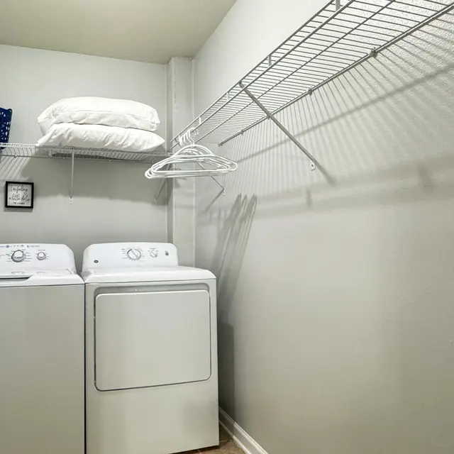 A compact laundry room featuring a washer and dryer side by side, with a wire shelf above holding white pillows and a blue basket. There is a small framed picture on the wall and the room is painted in a light gray color.