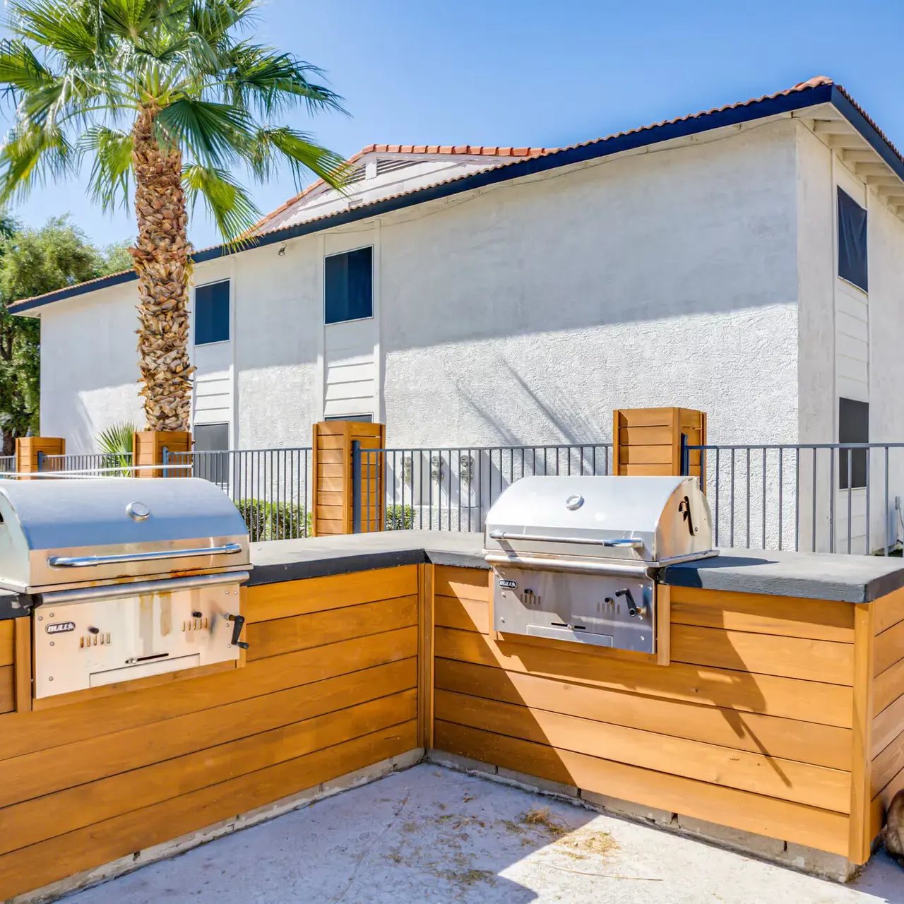 Outdoor grilling area featuring two stainless steel grills surrounded by wooden structures and a palm tree nearby.