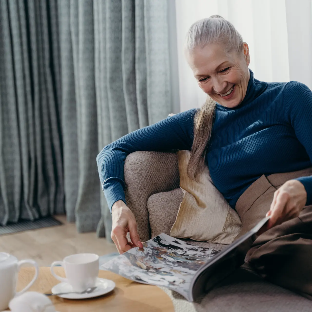 A woman with long hair, wearing a blue turtleneck sweater, sits comfortably on a sofa, smiling as she reads a magazine. A cup of tea and a sugar bowl sit on a wooden table in front of her.