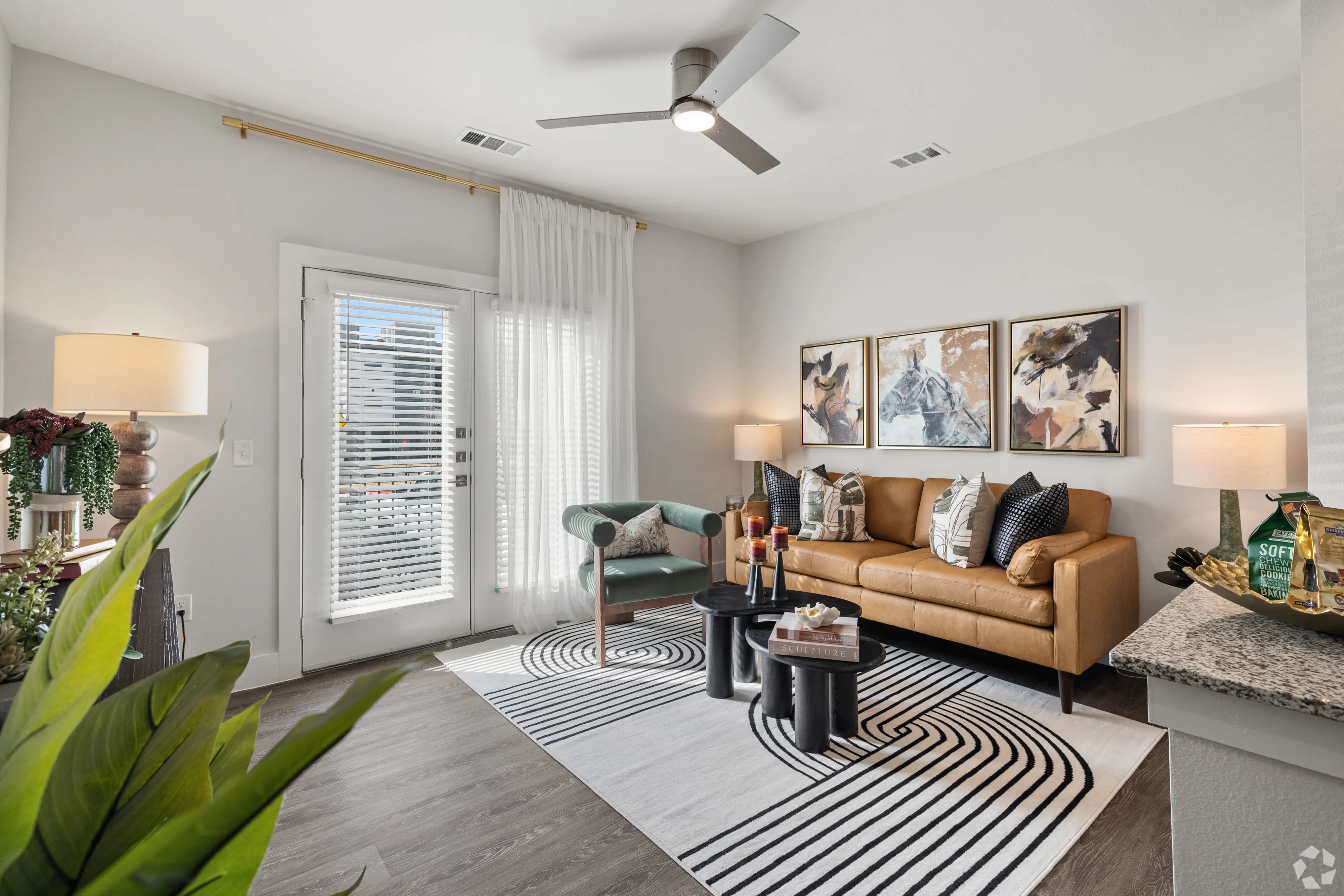 A modern living room featuring a brown leather sofa, a green armchair, a patterned area rug, and large windows with natural light. Decorative art pieces adorn the walls, and potted plants add a touch of greenery.
