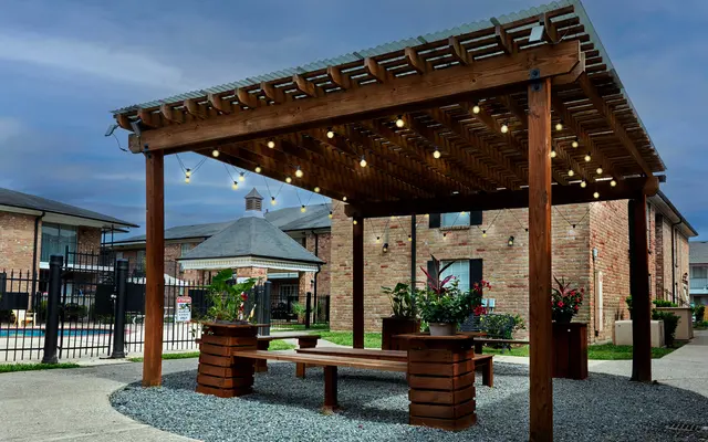An outdoor pavilion with wooden beams, string lights, and benches surrounded by plants, near a brick building and swimming pool area.