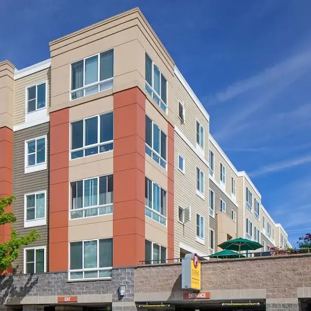 A modern, four-story apartment building featuring a mix of beige and orange facade, large windows, and a blue sky in the background. Surrounding greenery adds to the atmosphere.