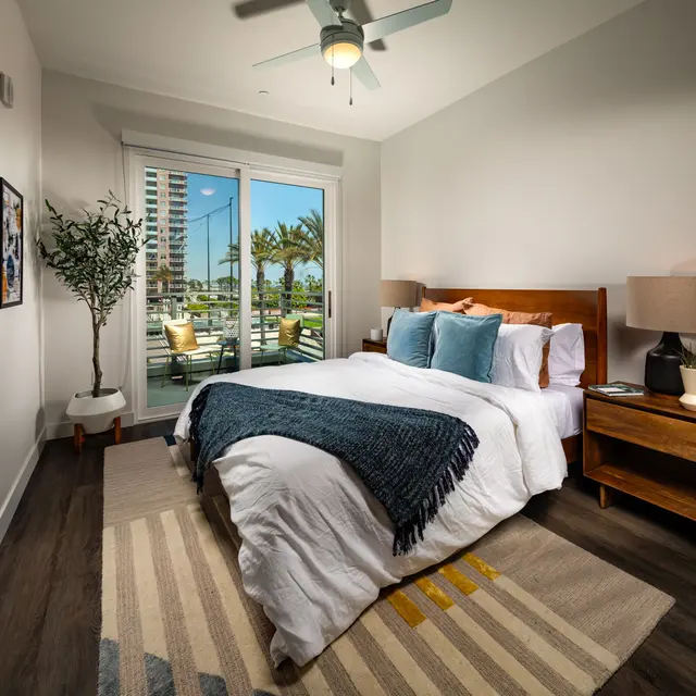 Modern Bedroom with Scenic View A well-decorated modern bedroom featuring a large bed with white linens and turquoise pillows. A wooden nightstand holds stylish lamps, and a decorative rug lies on the floor. Large windows overlook a patio and palm trees, offering a bright and inviting atmosphere.