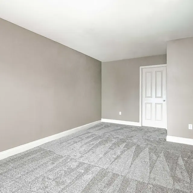 A spacious empty room with light gray walls and a carpeted floor. A closed white door is visible on the right side.