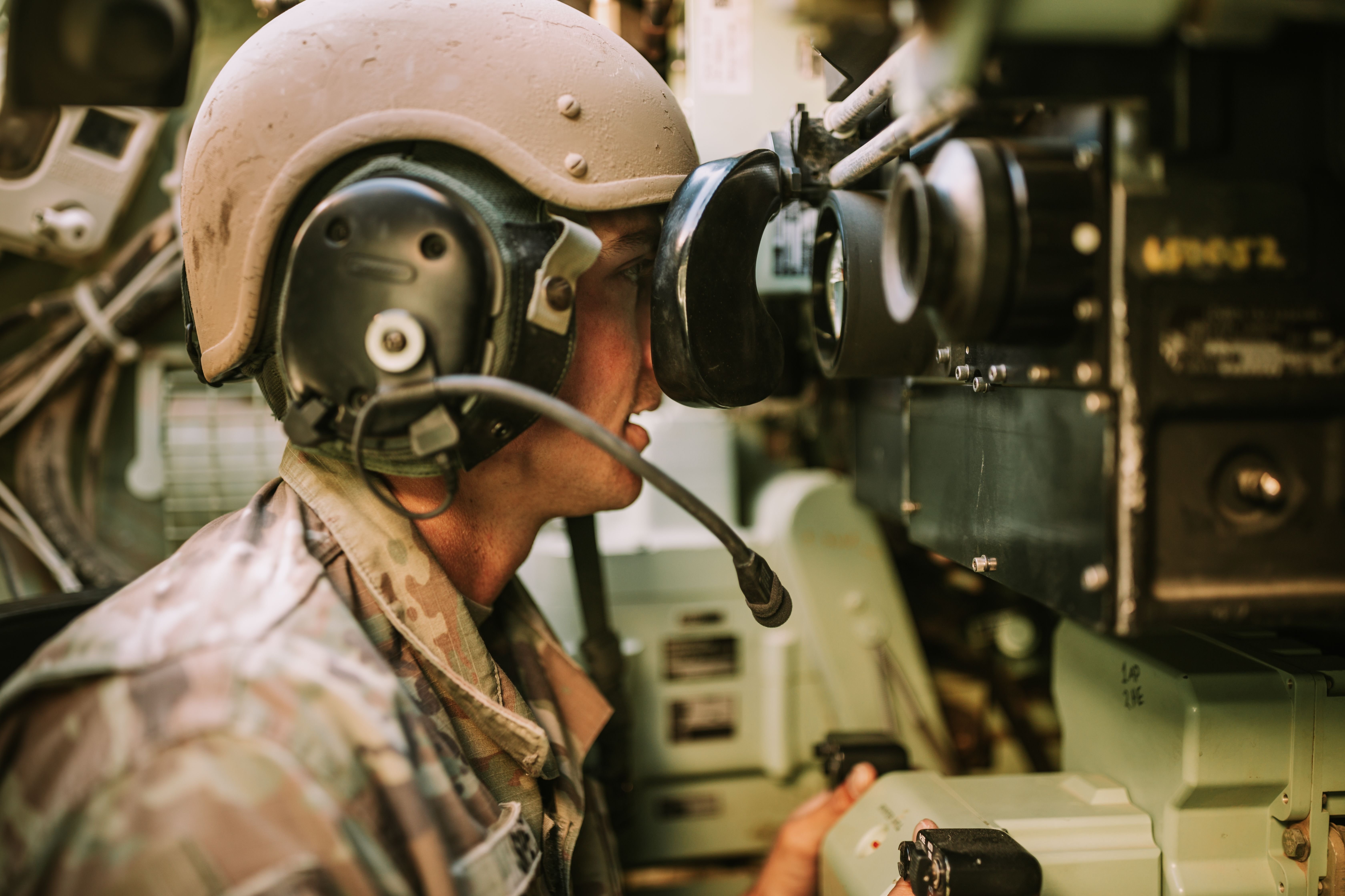 A soldier wearing a combat helmet and headphones observes through a military device, focusing intently on something in front of him.