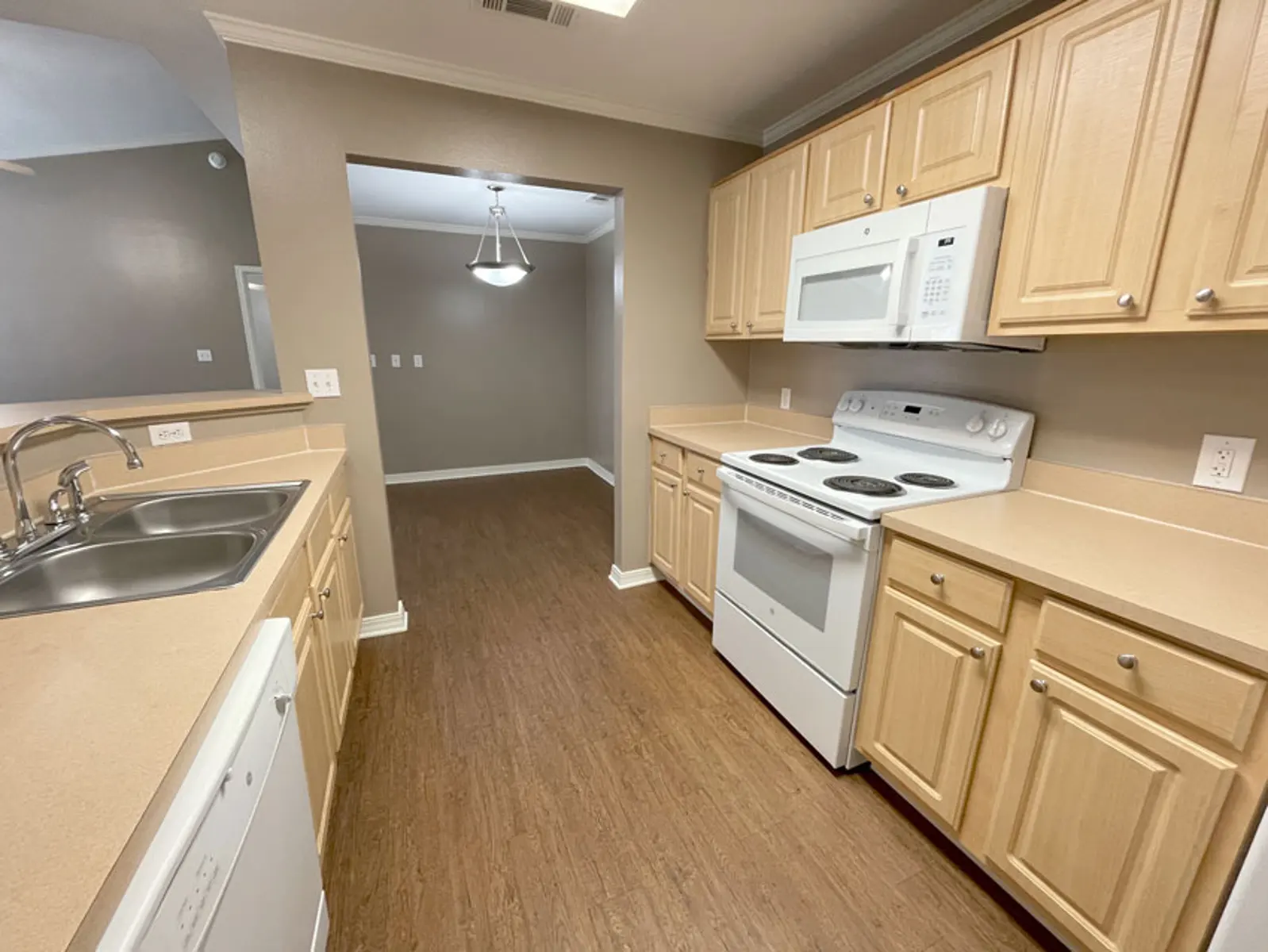 A modern kitchen with light brown cabinets and white appliances, featuring a dishwasher, stove, and microwave, with a brown laminate floor.