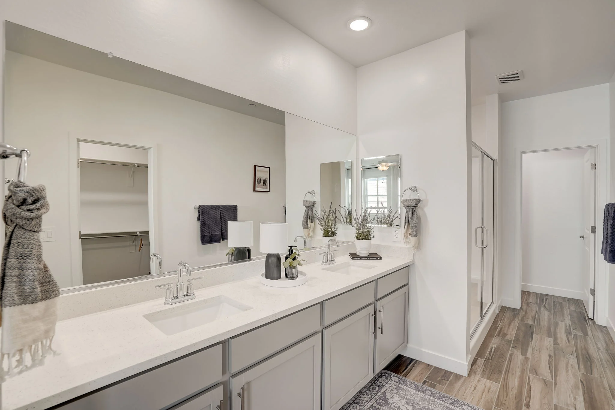 A modern bathroom featuring a double vanity with a light-colored countertop, two sinks, stylish faucets, and large mirrors. The bathroom includes a glass shower enclosure, wooden flooring, and light-colored walls with decorative plants and towels.