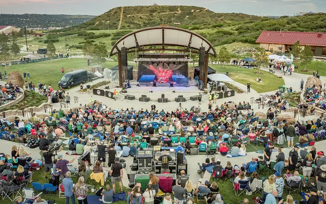A large crowd gathered for an outdoor concert at a spacious venue, featuring a stage with lights and equipment, surrounded by green hills and open space.