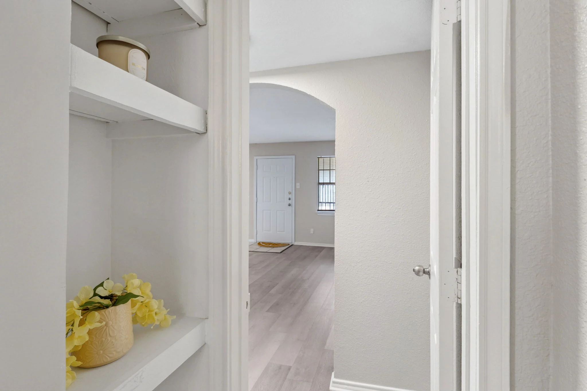 Entrance View of an Apartment A view from a hallway showing a shelf with decorative items and a doorway leading to a living space with light-colored flooring.