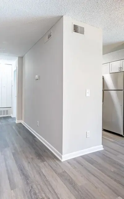 Interior view of a modern, spacious apartment featuring light wooden floors, neutral wall colors, and a partial view of a kitchen with a refrigerator.