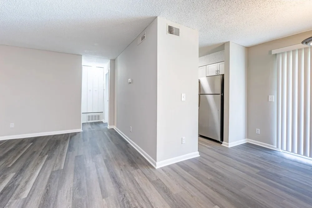 Interior view of a modern, spacious apartment featuring light wooden floors, neutral wall colors, and a partial view of a kitchen with a refrigerator.