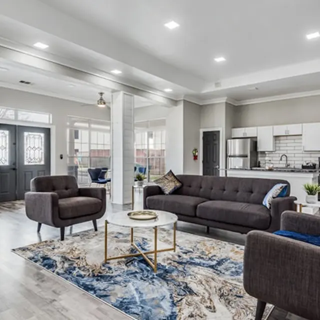 A stylish modern living room featuring a gray sofa, armchairs, and a round coffee table on a decorative rug. The space has an open layout with a kitchen visible in the background, equipped with modern appliances. Natural light floods the room through large windows.