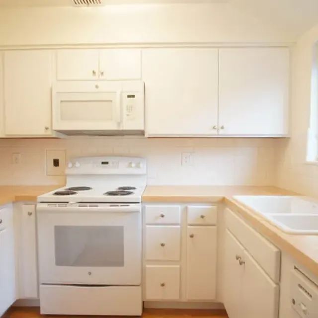 A modern kitchen featuring white cabinets and appliances, including a refrigerator, microwave, and stove, with a wooden countertop and a double sink near a window.