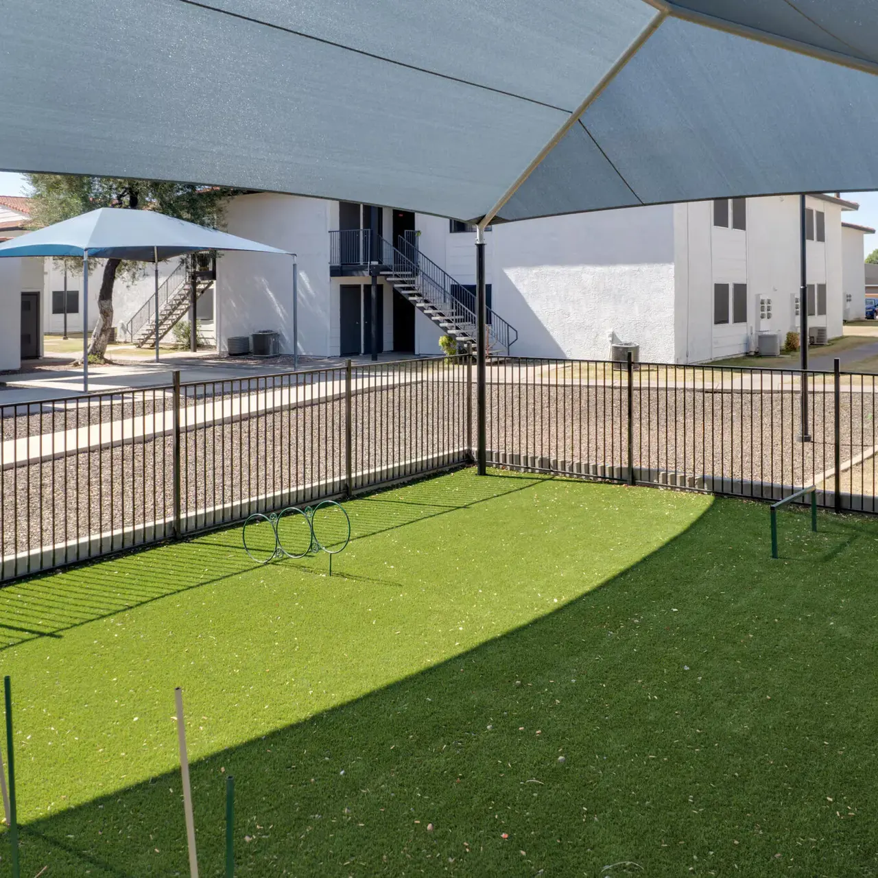 A fenced dog park area featuring artificial grass and agility equipment, under a shade structure.