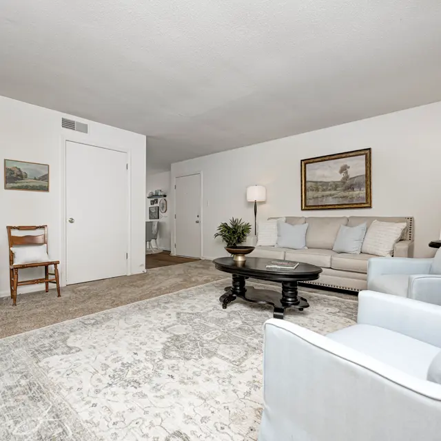 A spacious living room featuring a light color scheme, a comfortable sofa, a couple of armchairs, a round coffee table, and a bookshelf. The decor is simple and elegant, with a painting on the wall and a small plant on the coffee table.
