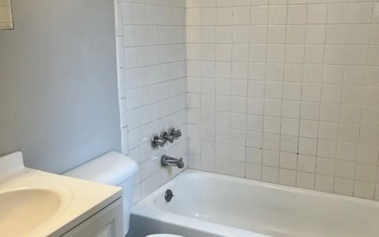 A small, simple bathroom featuring a bathtub with a white tiled surround, a showerhead, a toilet, and a vanity with a sink. The walls are painted light gray and the floor features small square tiles.
